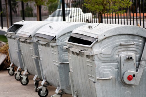 Four large, industrial metal wheelie bins lined up on a paved surface, likely part of a commercial rubbish collection service near Wandsworth. The bins are constructed from galvanized steel with a textured, slightly reflective surface, showing signs of weathering and use. Each bin has a curved, hinged lid with a black handle, and the lids are closed, with one slightly open at an angle. The bins are positioned closely side by side, with their small, durable wheels visible at the front, allowing for easy movement. In the background, a black metal fence and some green foliage can be seen, along with parked vehicles on a street, suggesting an urban or suburban setting. The overall scene appears to be outdoors during daylight, with natural lighting illuminating the metallic surfaces. This image relates to commercial waste removal services in Wandsworth, where reputable companies such as Commercial Waste Wandsworth manage rubbish collection efficiently.