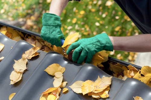 A person wearing green gardening gloves is manually removing fallen yellow and brown leaves from a dark grey plastic guttering system installed on the edge of a building roof, with dense foliage and green shrubbery visible in the blurred background. The leaves, scattered across the gutter, vary in shape and size, indicating autumnal debris. The gutter appears clean but needs clearing to ensure proper drainage. The scene suggests routine outdoor maintenance, possibly conducted by a waste management or rubbish removal service in Wandsworth. The natural outdoor environment and the focus on debris removal highlight the importance of regular gutter clearance as part of property maintenance. The lighting is natural, indicating daylight conditions, with the contrast between the vibrant leaves and the darker gutter surface clearly visible. This image captures a typical activity associated with garden waste clearance and routine rubbish management in a residential or commercial setting.