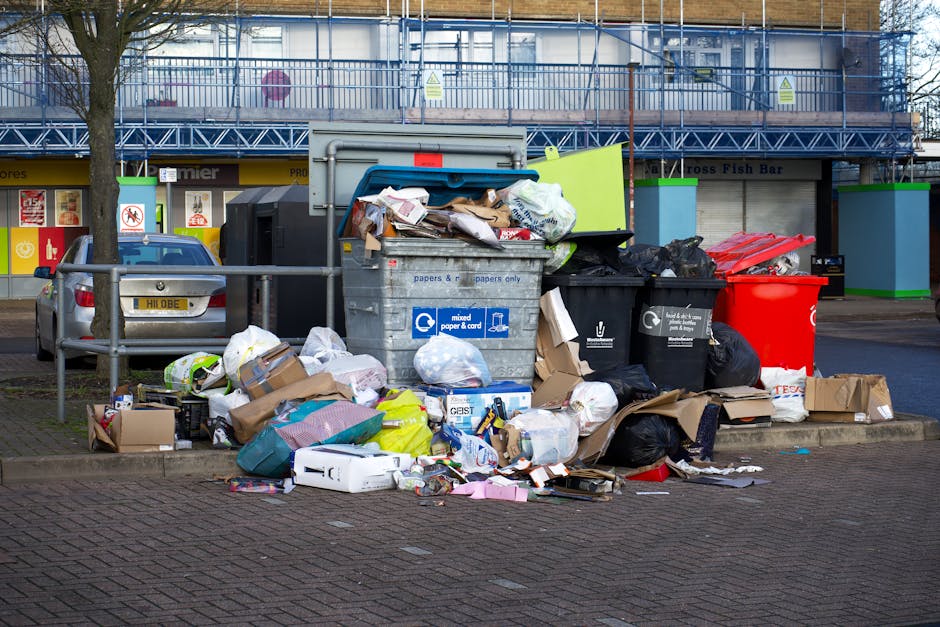 An aerial view of a large amount of mixed waste and rubbish on the ground in an open area, including scattered plastic bags, cardboard boxes, and various discarded household items. In the center, thre