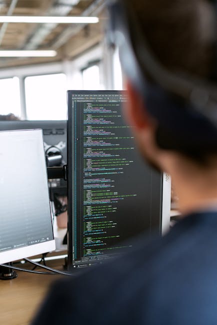 A person’s hand operating a black computer keyboard on a white desk, with a large monitor screen displaying lines of code or text in the background. The workspace includes a white laptop partially v