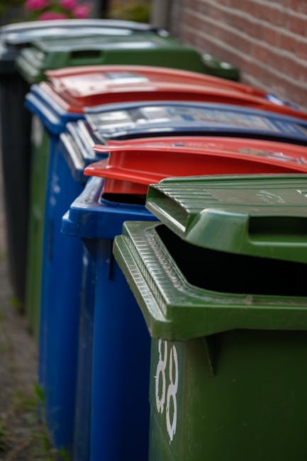 A row of large plastic wheelie bins lined up along a pavement beside a brick wall, with colours including blue, green, red, and dark green. Each bin features a flat, rectangular lid with a ribbed text